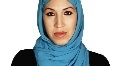 An image of a Brown woman wearing a blue hijab and a black long sleeved shirt, looking straight at camera. 